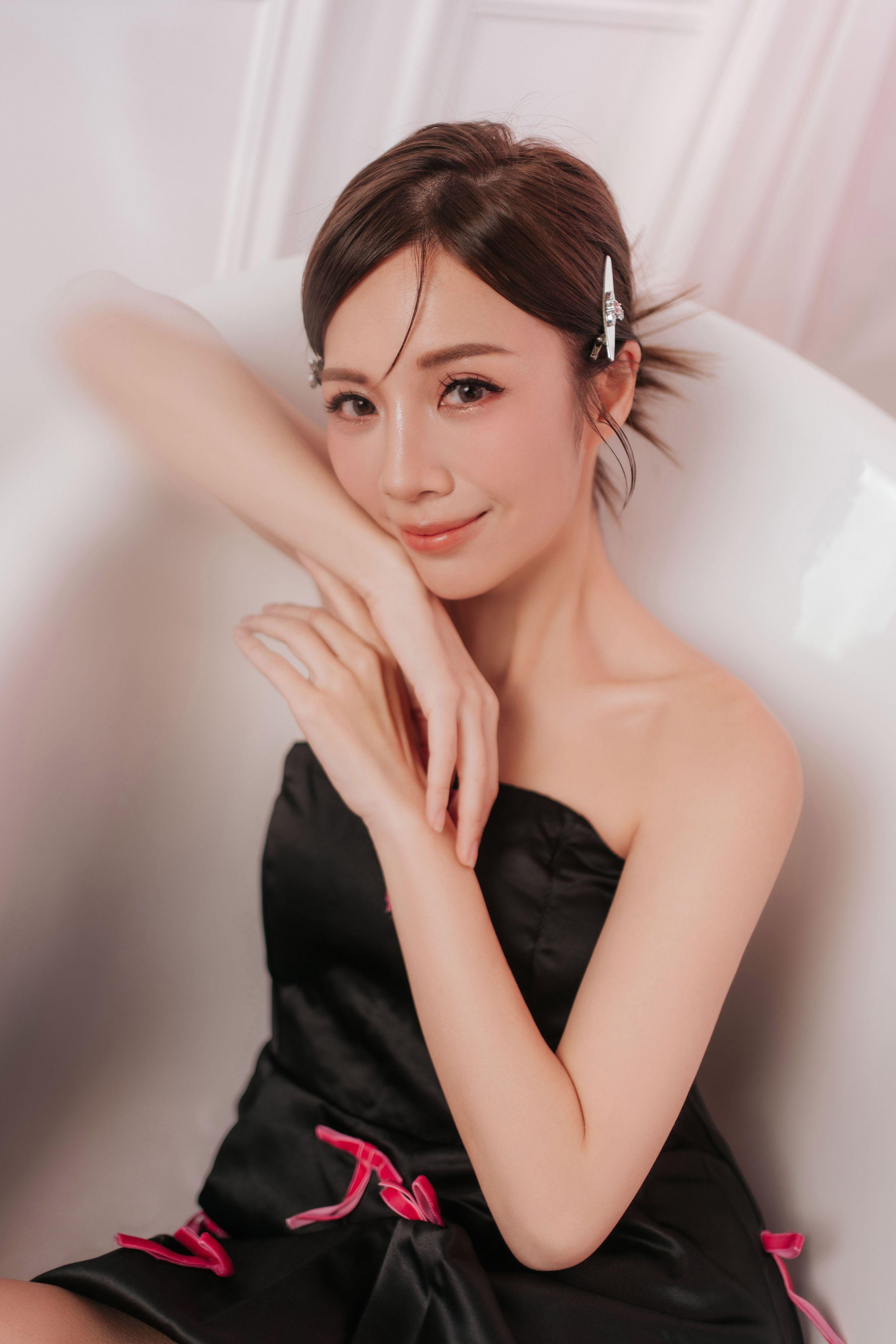 香港藝人賴彥妤自然神情特寫Young Asian woman in black strapless dress with pink accents, hair up with pearl clip, posing gracefully on white chair.