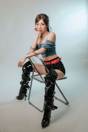 香港人像攝影作品，呈現自然與時尚感Young Asian woman in denim crop top, black shorts, red belt, and thigh-high patent boots seated on folding chair.
