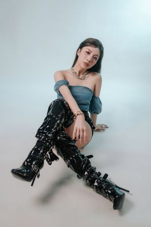 香港藝人賴彥妤 Vivienna Lai 形象照Young woman in denim off-shoulder top, patent black thigh-high boots, and chain jewelry poses on white studio floor.