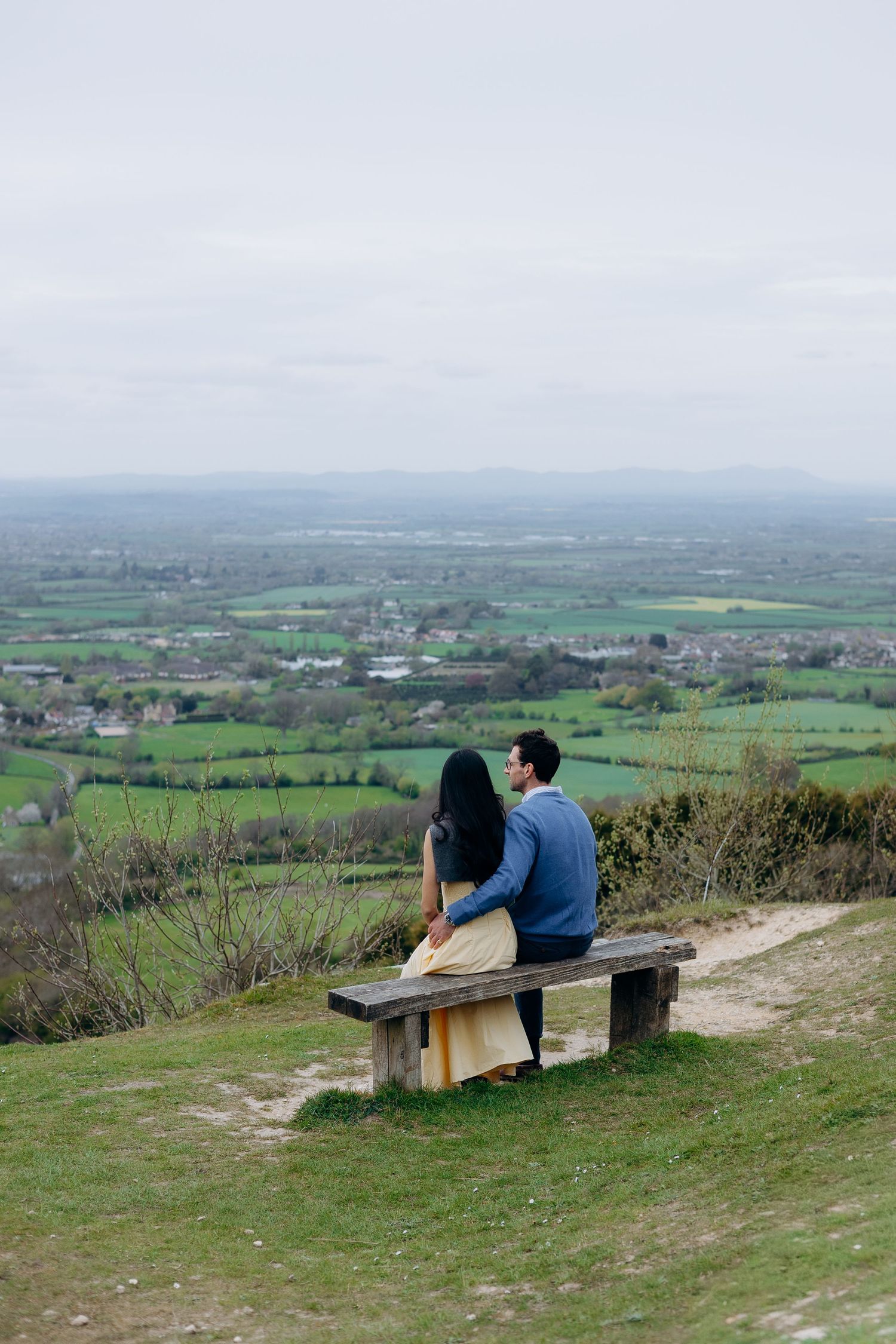 Couple sitting on a wooden bench on a hilltop overlooking a vast green countryside landscape on a cloudy day.