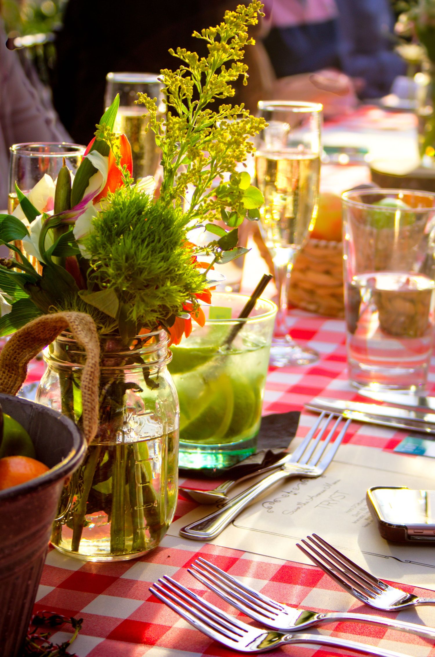 Outdoor dining table set with red checkered cloth, fresh wildflowers in mason jar vase, and glassware in warm sunlight.