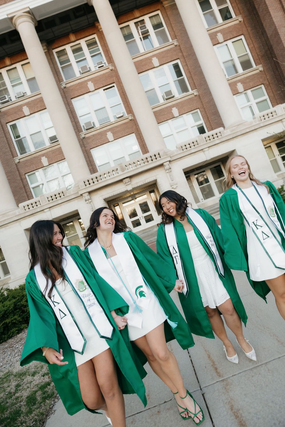 Four laughing Kappa Delta graduates in green robes and white dresses celebrate outside a brick university building.