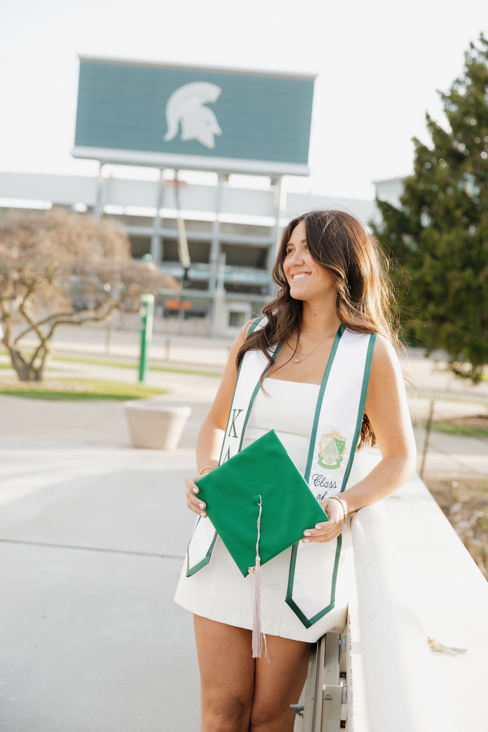 Brunette woman in white dress holding green MSU graduation cap near Spartan logo sign outdoors.