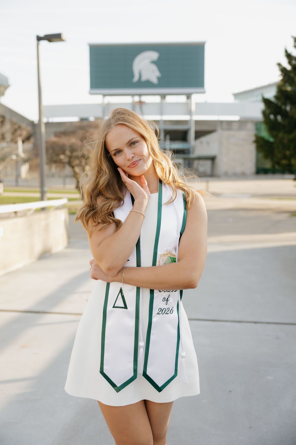 Blonde woman in white dress with Delta and Class of 2024 graduation stoles near Michigan State sign.