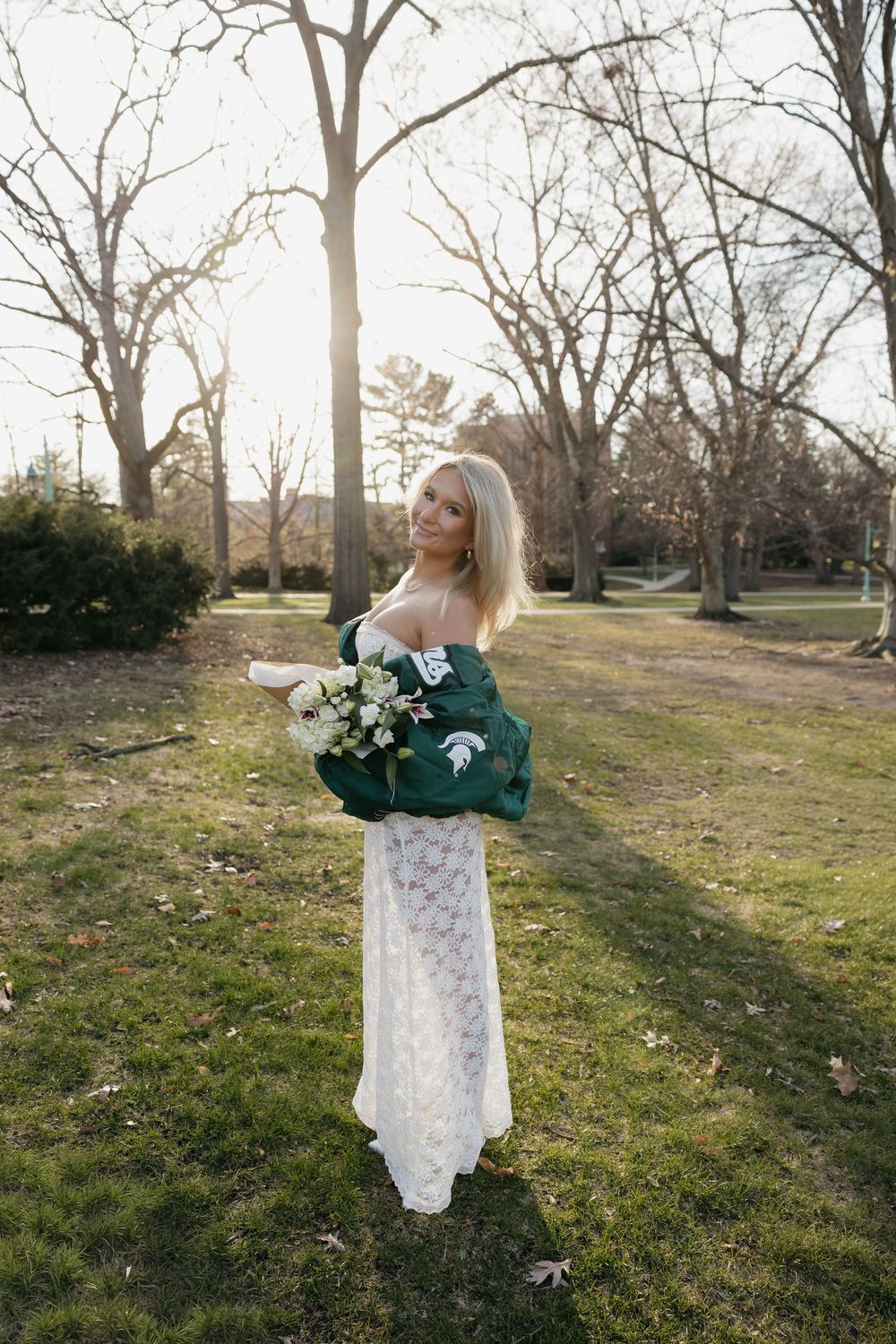 Blonde graduate in white lace dress holds green MSU jacket and white flowers in a sunlit campus park.