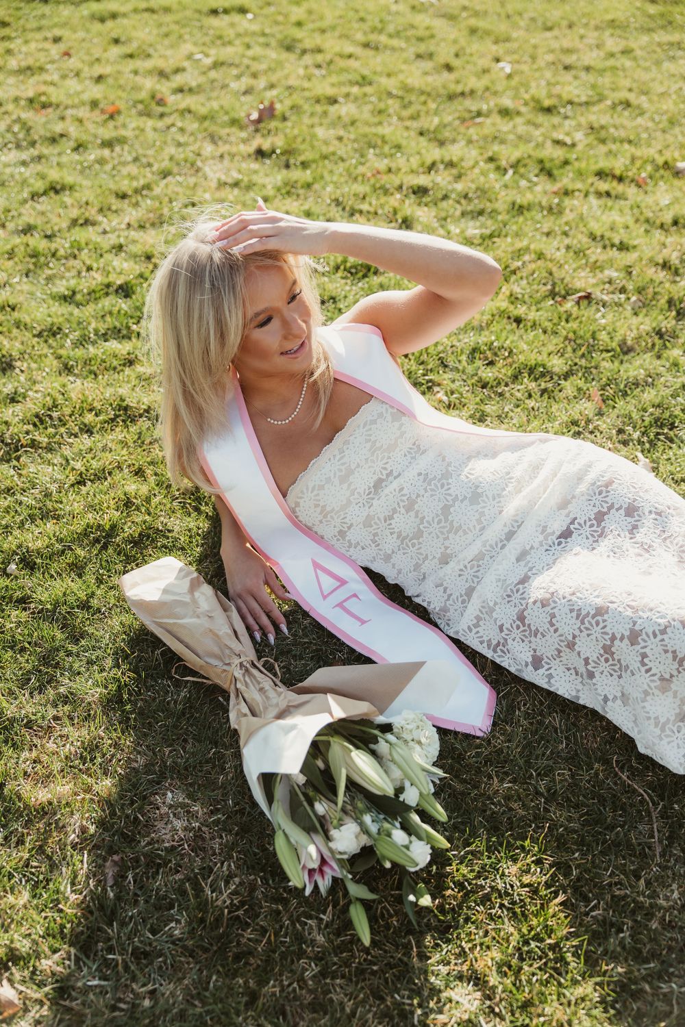 Blonde graduate in white lace dress and Delta Gamma stole reclines on grass holding white tulip bouquet.