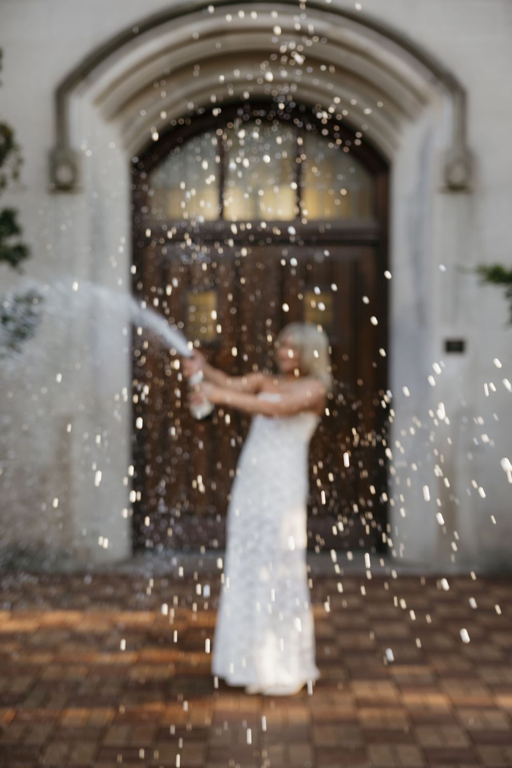 Graduate in white lace dress sprays champagne bottle in front of an arched stone building entrance.