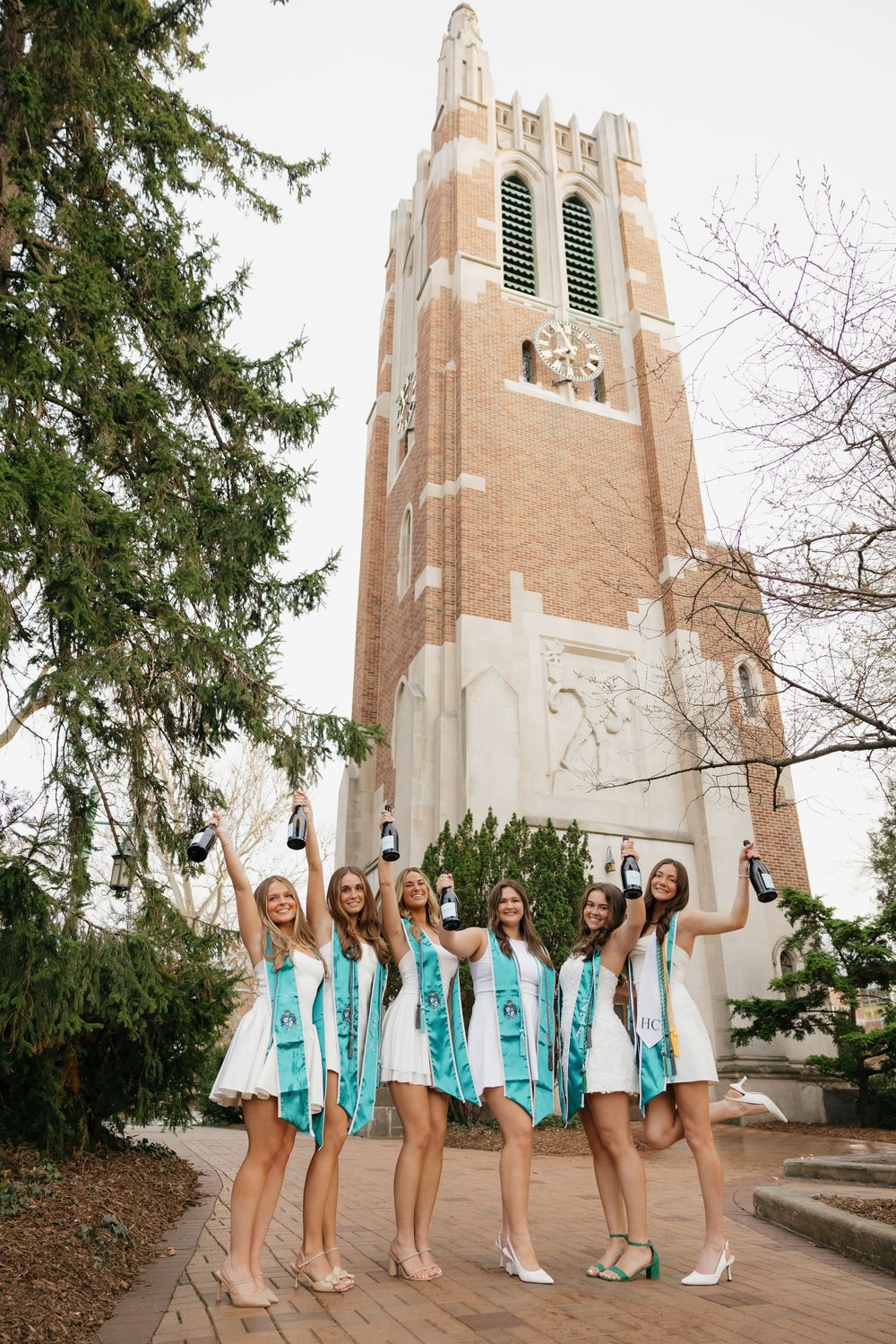 Group of six women in white dresses with teal graduation stoles celebrating near Beaumont Tower at MSU.