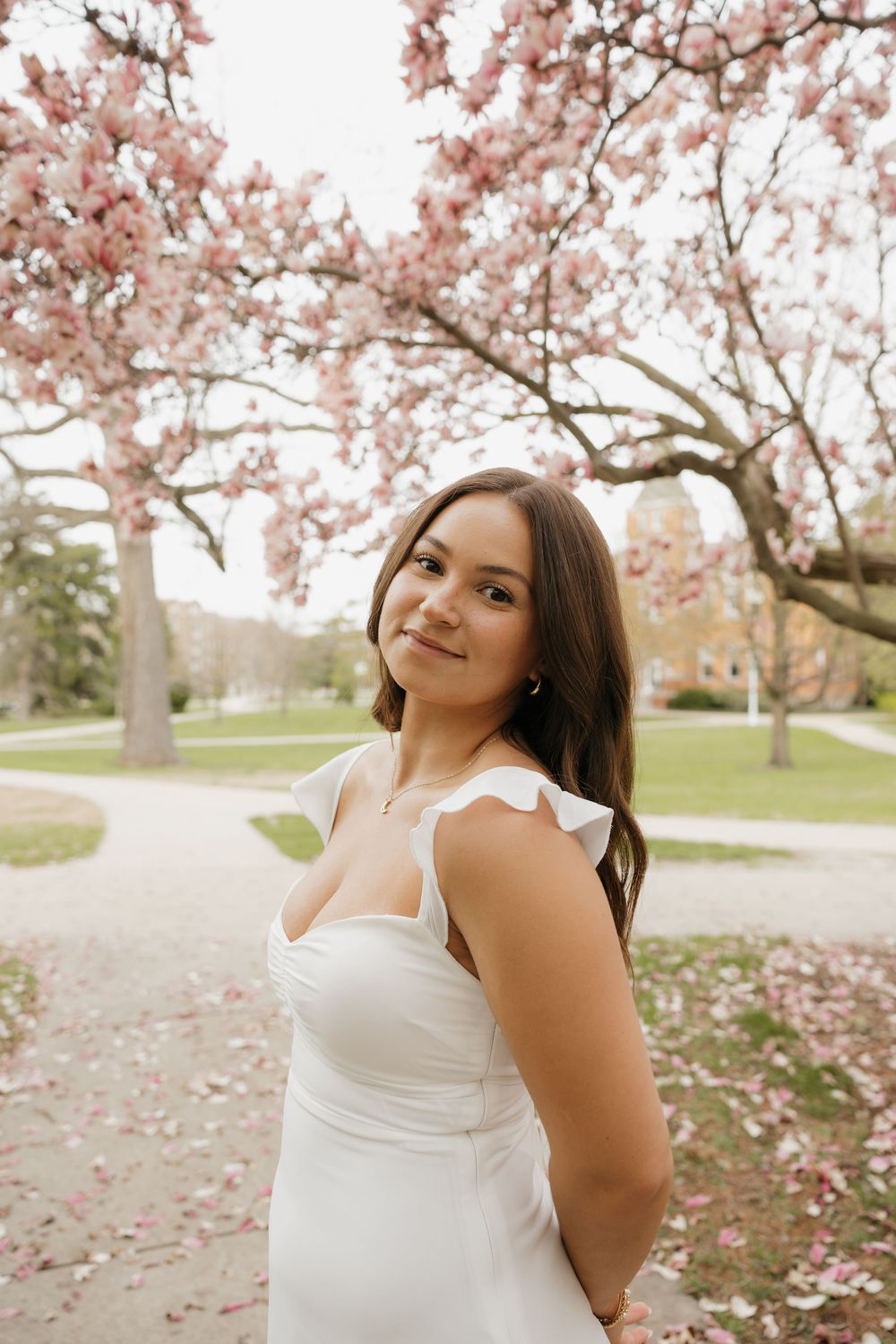 Brunette woman in white ruffled dress smiling under pink magnolia blossoms in a garden setting.