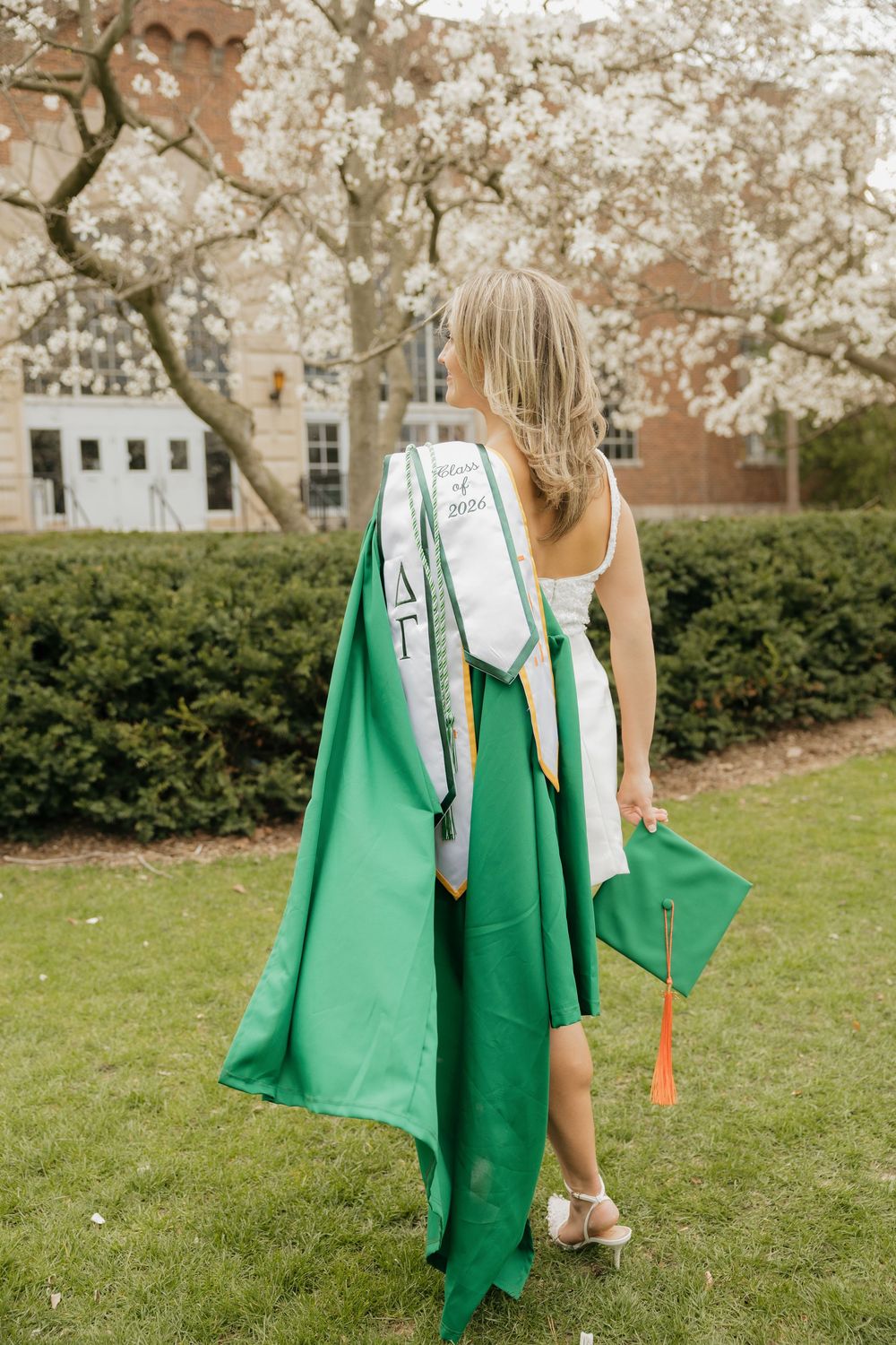 Blonde graduate from behind holding green cap and gown near flowering tree on campus.