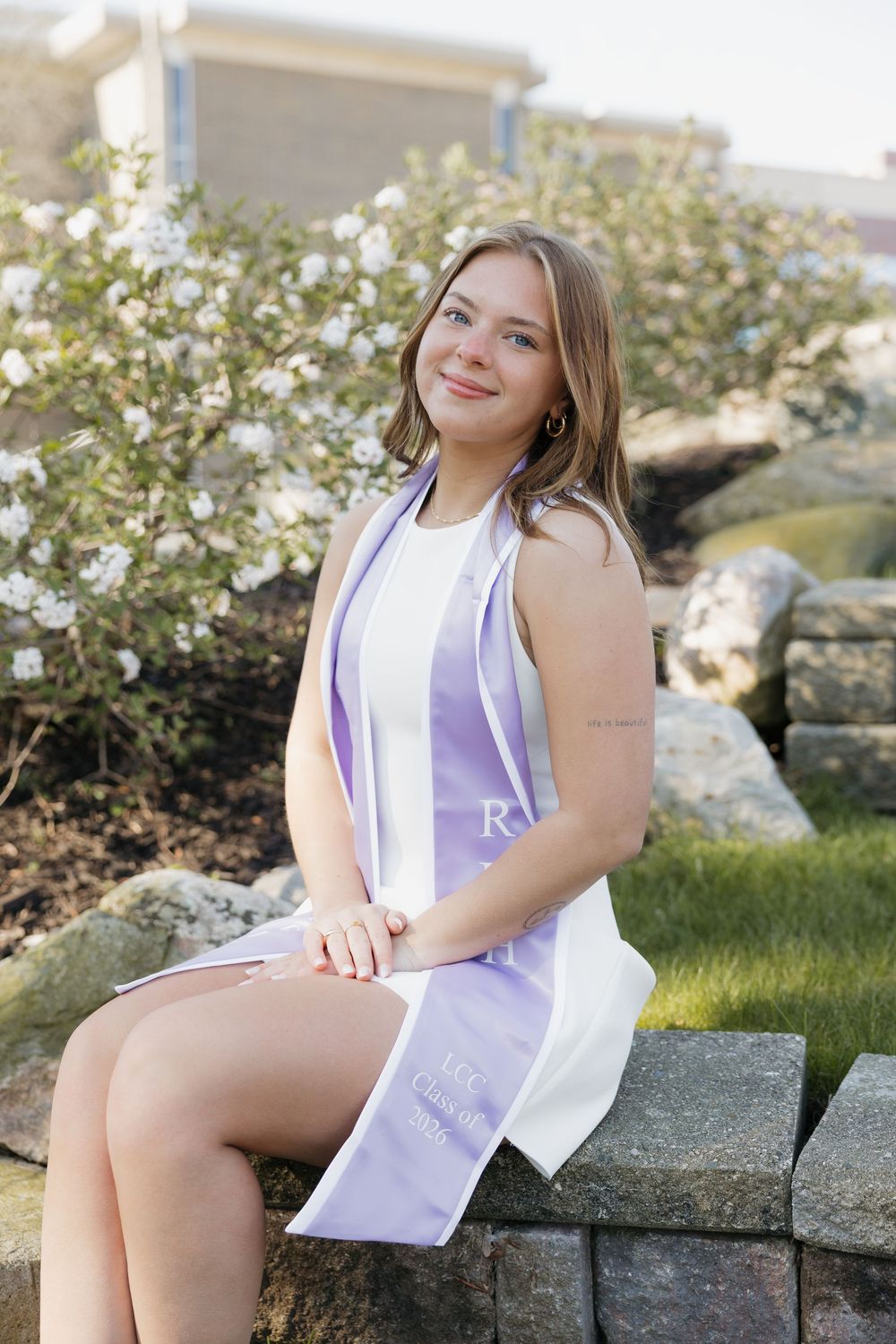 Smiling young woman in white dress with lavender graduation stole seated on stone wall.