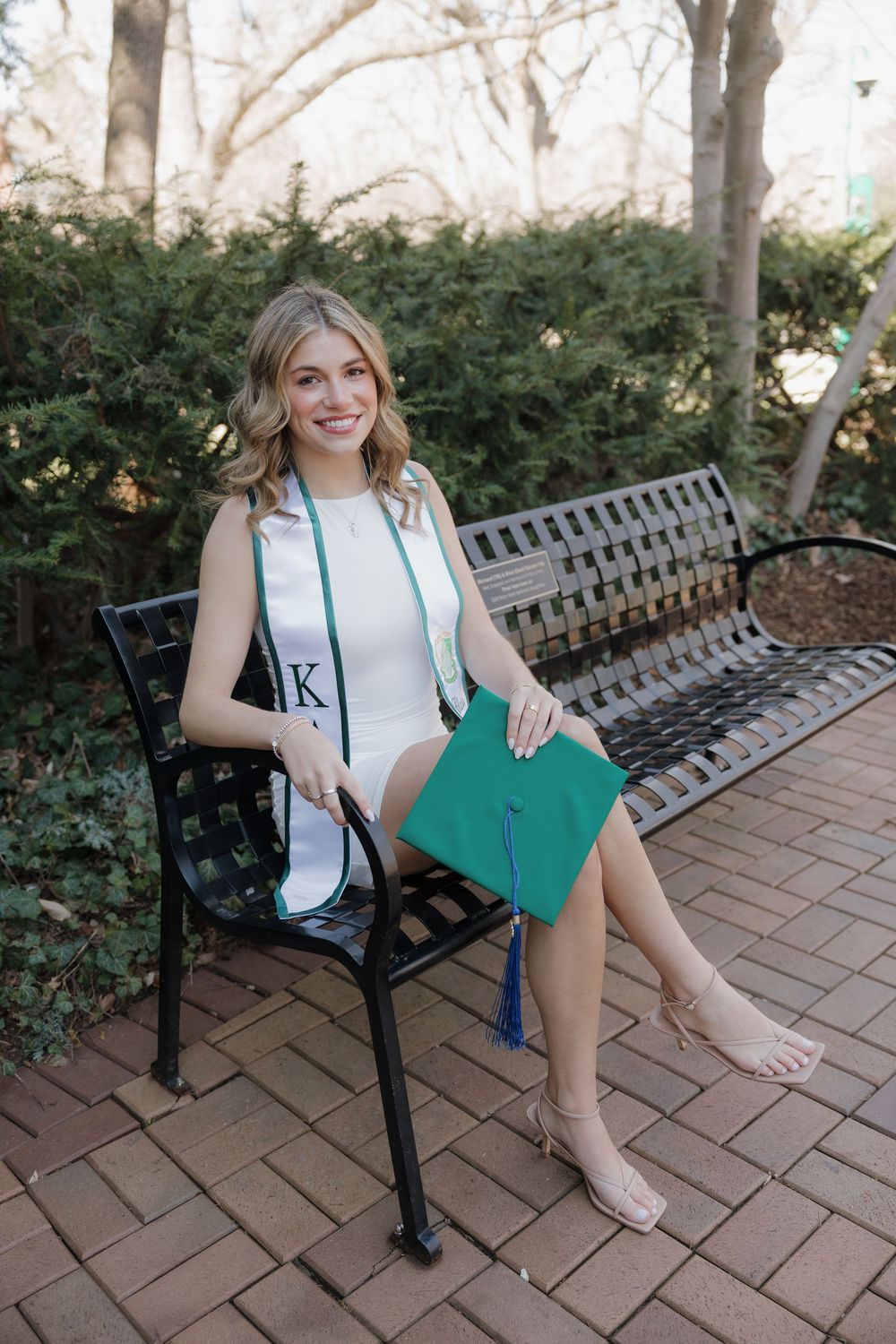 Smiling blonde woman in white dress holding green graduation cap while seated on a bench outdoors.