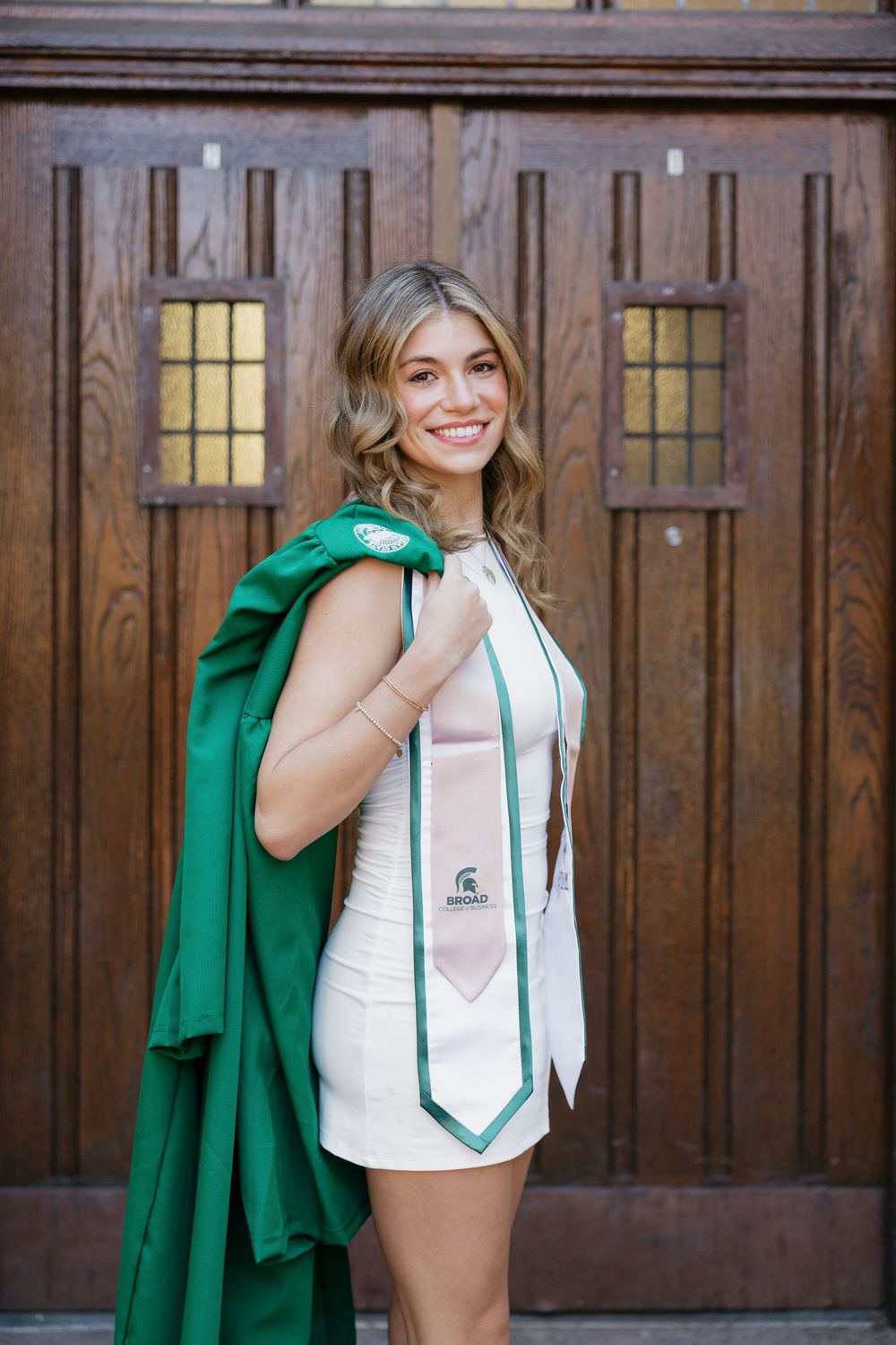 Smiling blonde woman in white dress holding green graduation gown over shoulder near wooden doors at MSU.
