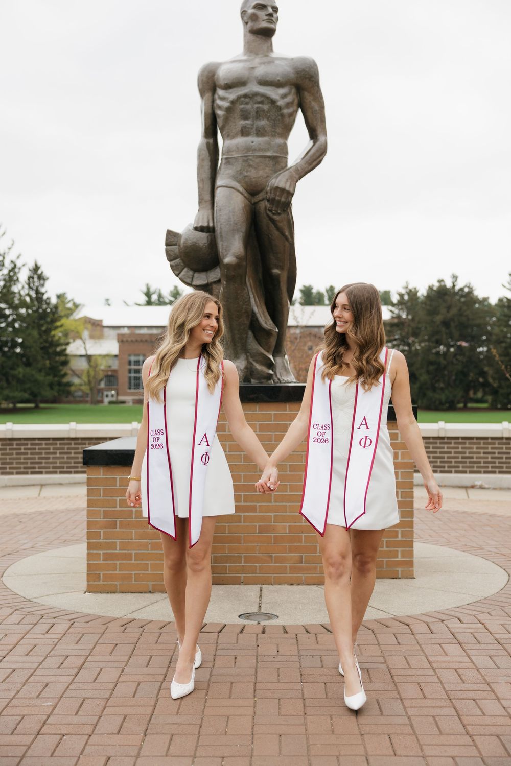 Two blonde graduates in white dresses with Alpha Phi stoles posing by Spartan statue.