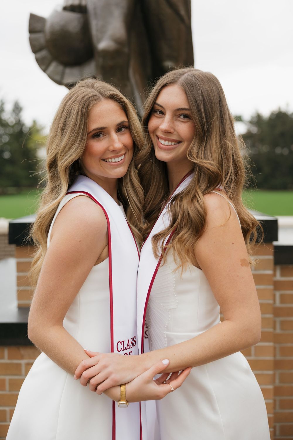 Two smiling female graduates in white dresses with red stoles posing by campus statue.
