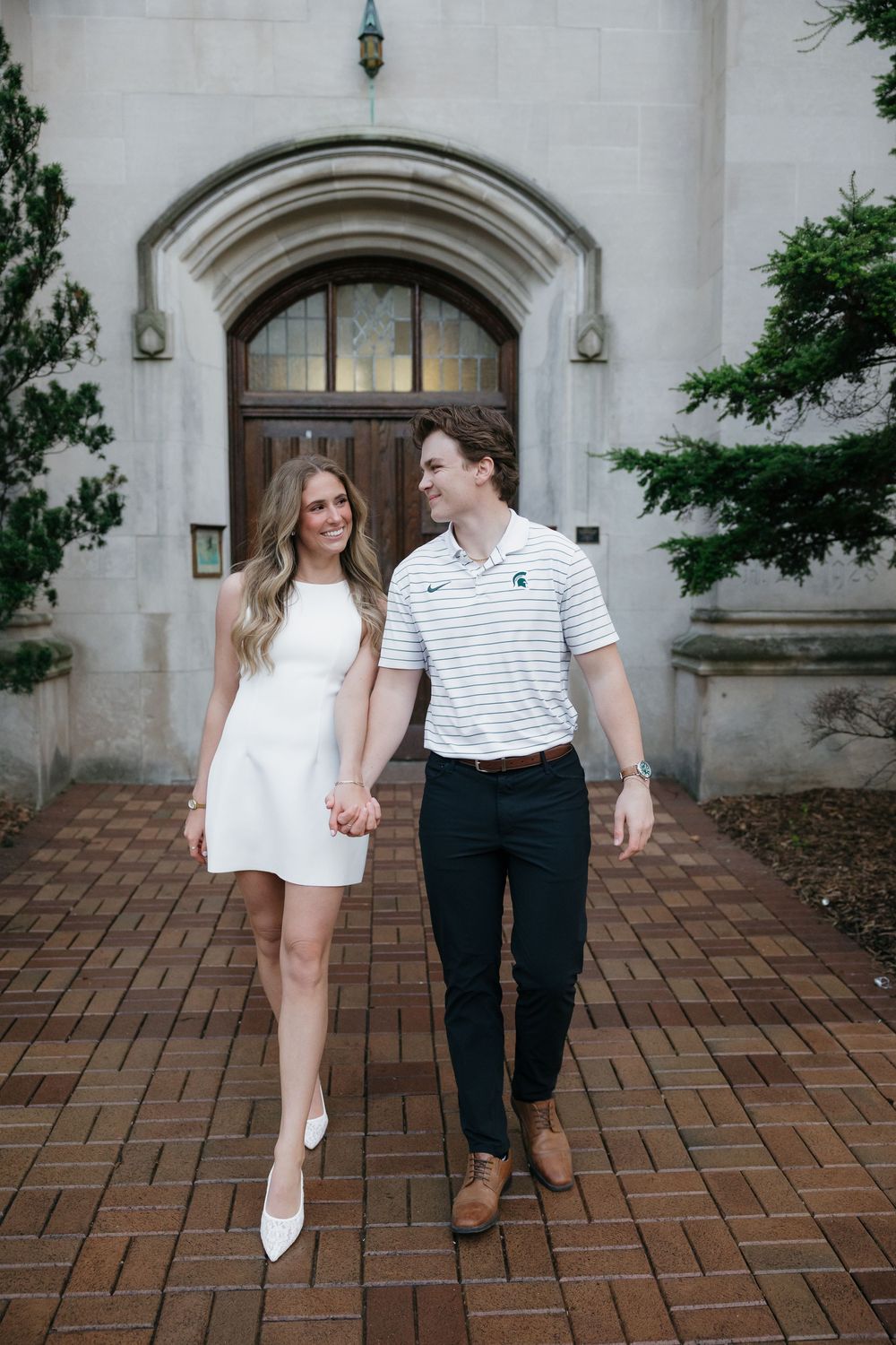 Smiling couple in white outfits walking hand in hand outside historic stone building.