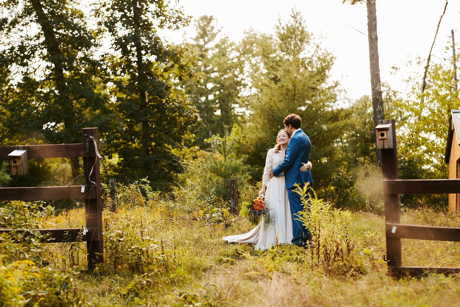 Couple embracing in sunlit field with rustic wooden fence.