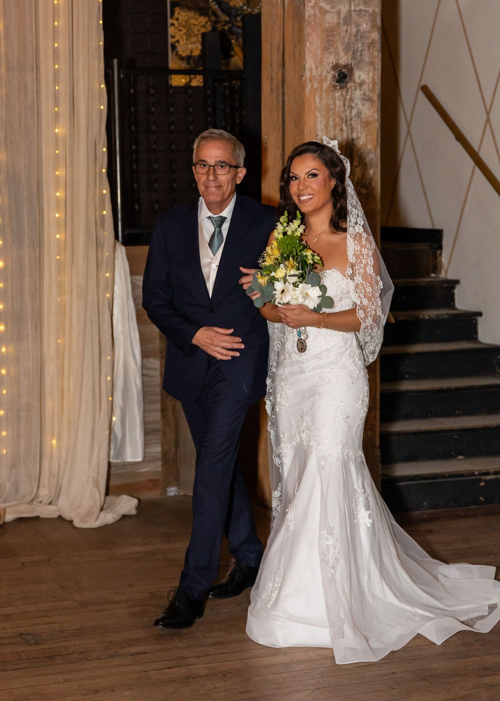 Bride in white lace mermaid gown holding yellow bouquet walks with older man in navy suit at elegant indoor wedding venue.