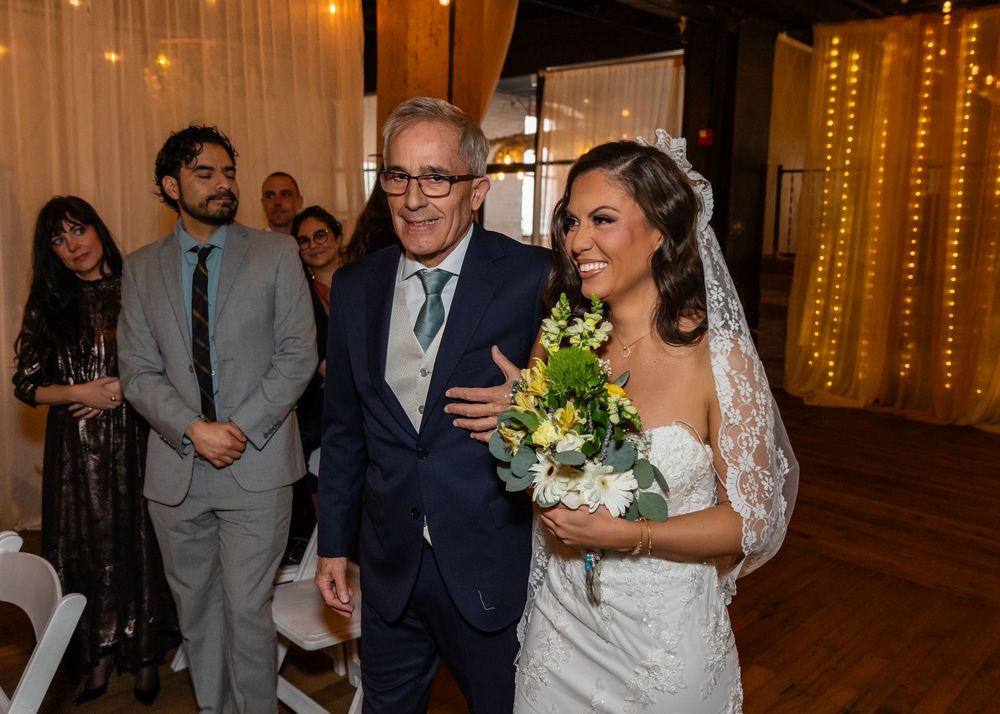 Bride in white gown and veil walks down aisle with older man in navy suit, holding yellow floral bouquet at indoor wedding.