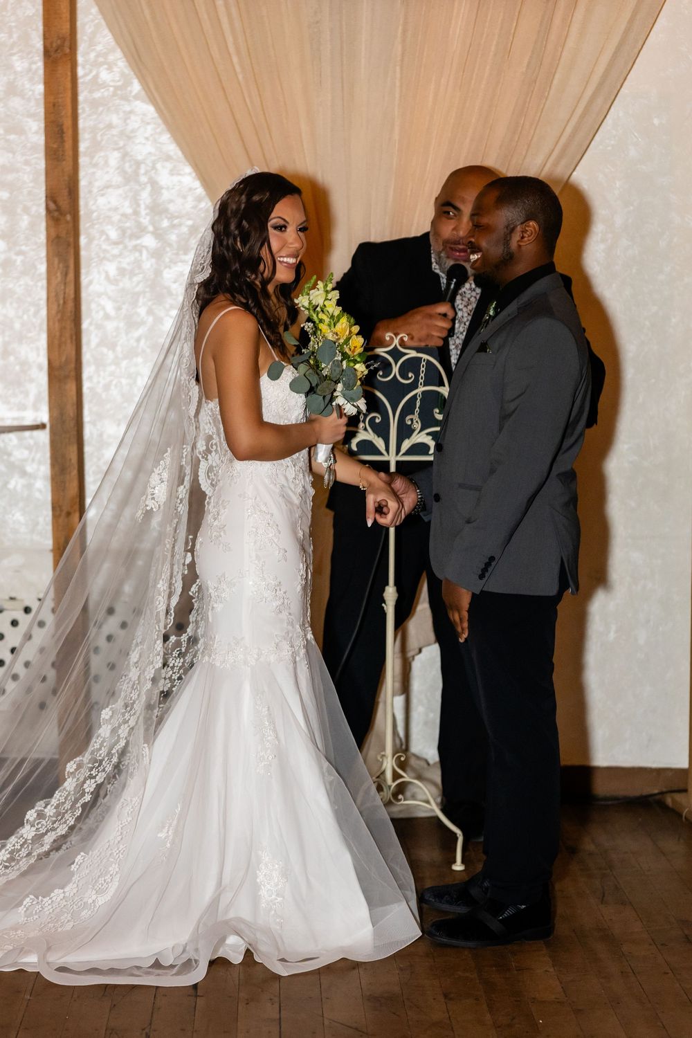 Bride in lace mermaid gown holds yellow bouquet as groom exchanges vows at elegant indoor wedding ceremony.