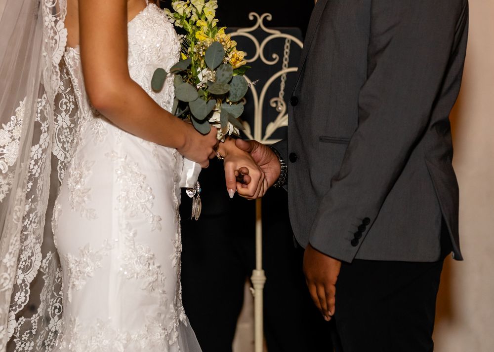 Bride and groom holding hands during wedding ceremony, bride wearing lace gown and veil holding greenery bouquet.