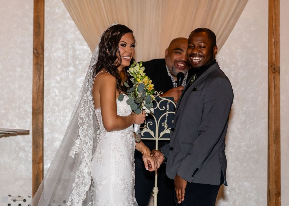 Smiling bride and groom celebrate with officiant during joyful wedding ceremony, bride in lace gown with veil.