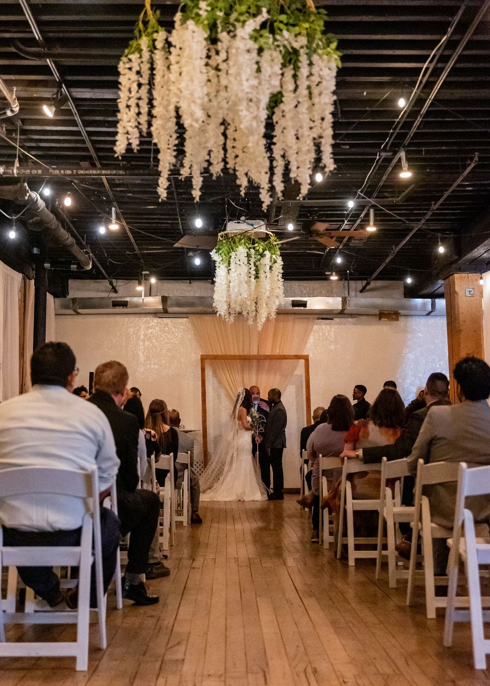 Indoor wedding ceremony with floral chandelier, white chairs, and couple at wooden arch altar.