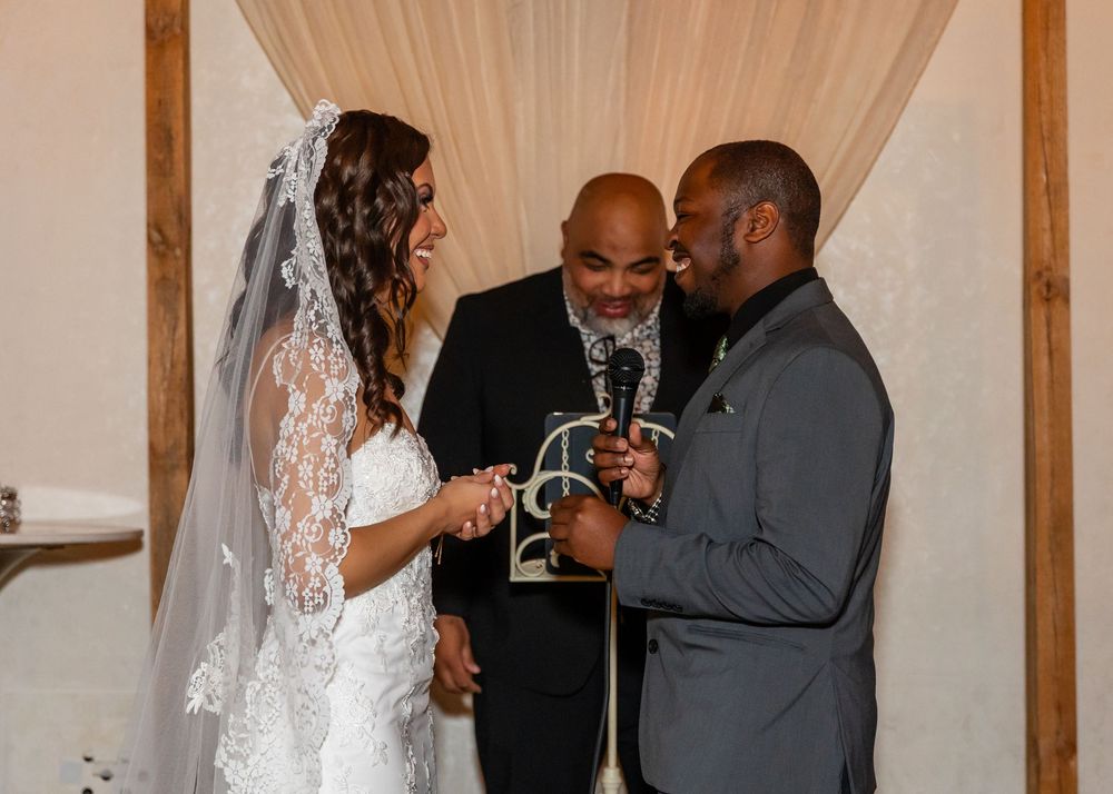 Smiling bride and groom share a joyful moment with officiant during indoor wedding ring exchange ceremony.