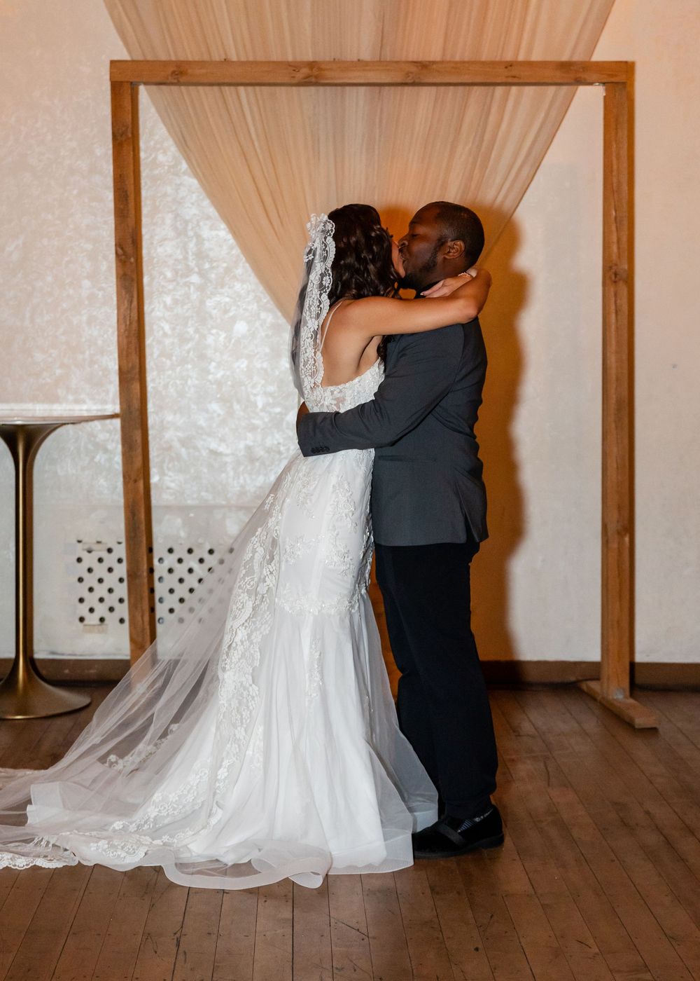 Newlyweds embrace in a passionate first kiss under a rustic wooden arch at their wedding ceremony.