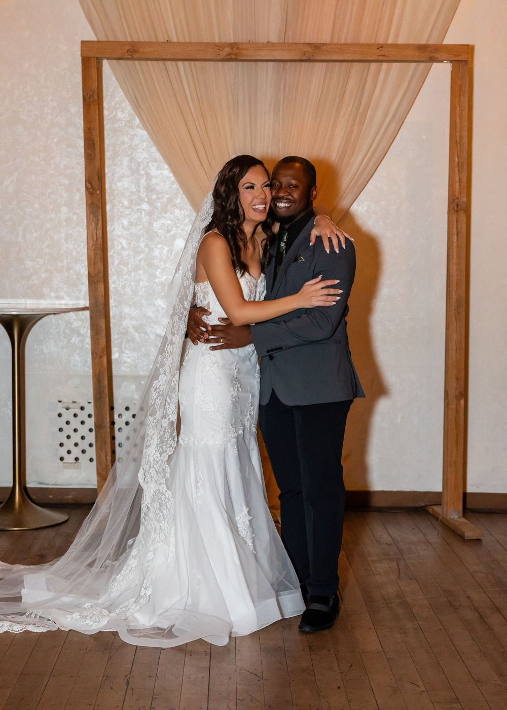 Smiling bride in lace gown and groom in suit embrace before wooden arch with draped fabric.