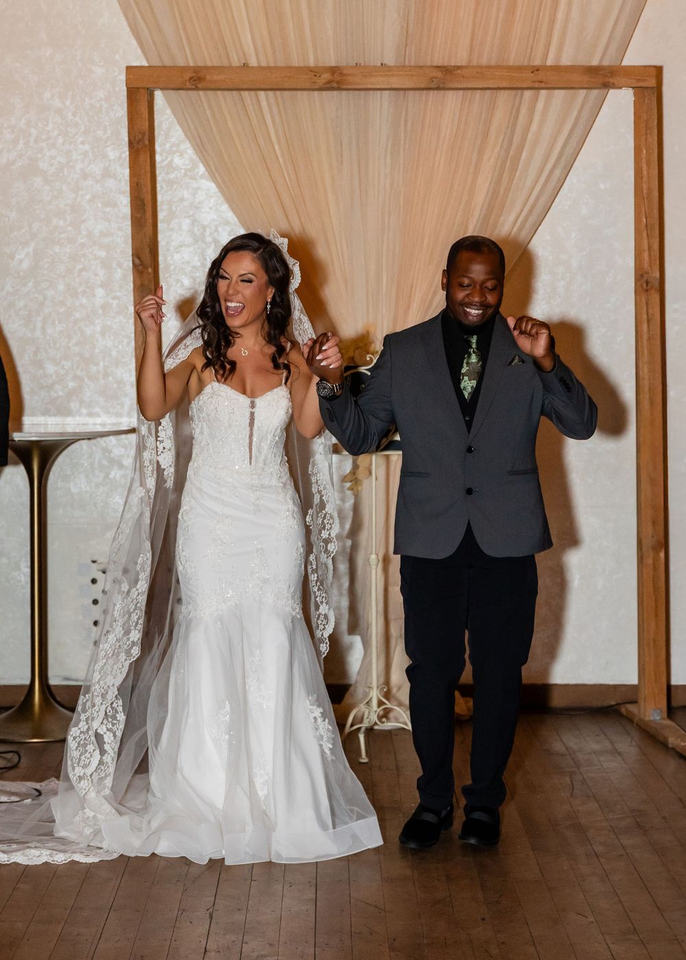Jubilant newlyweds celebrate under wooden arch with flowing drapes at elegant indoor wedding reception.