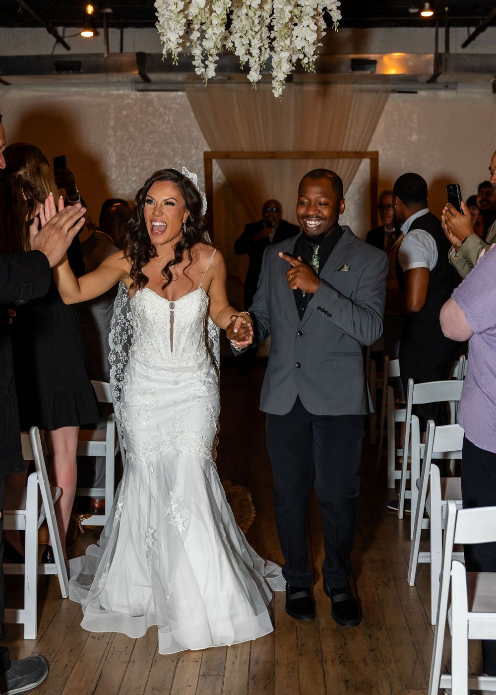 Radiant newlyweds walk down aisle as guests applaud at elegant indoor wedding ceremony celebration.