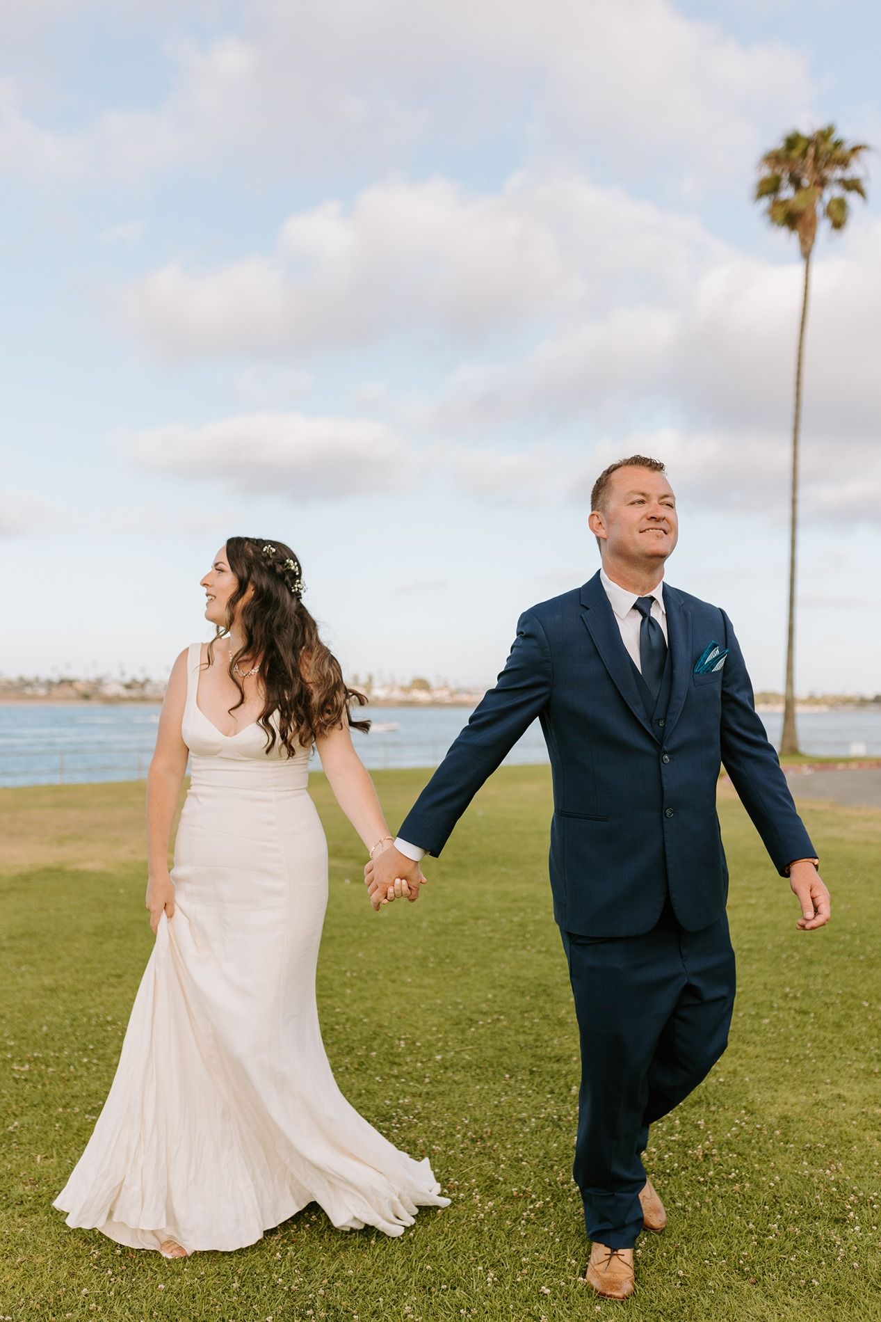 Bride and groom walk hand-in-hand on green lawn near waterfront, palm tree and cloudy sky backdrop at Tower Beach Club.