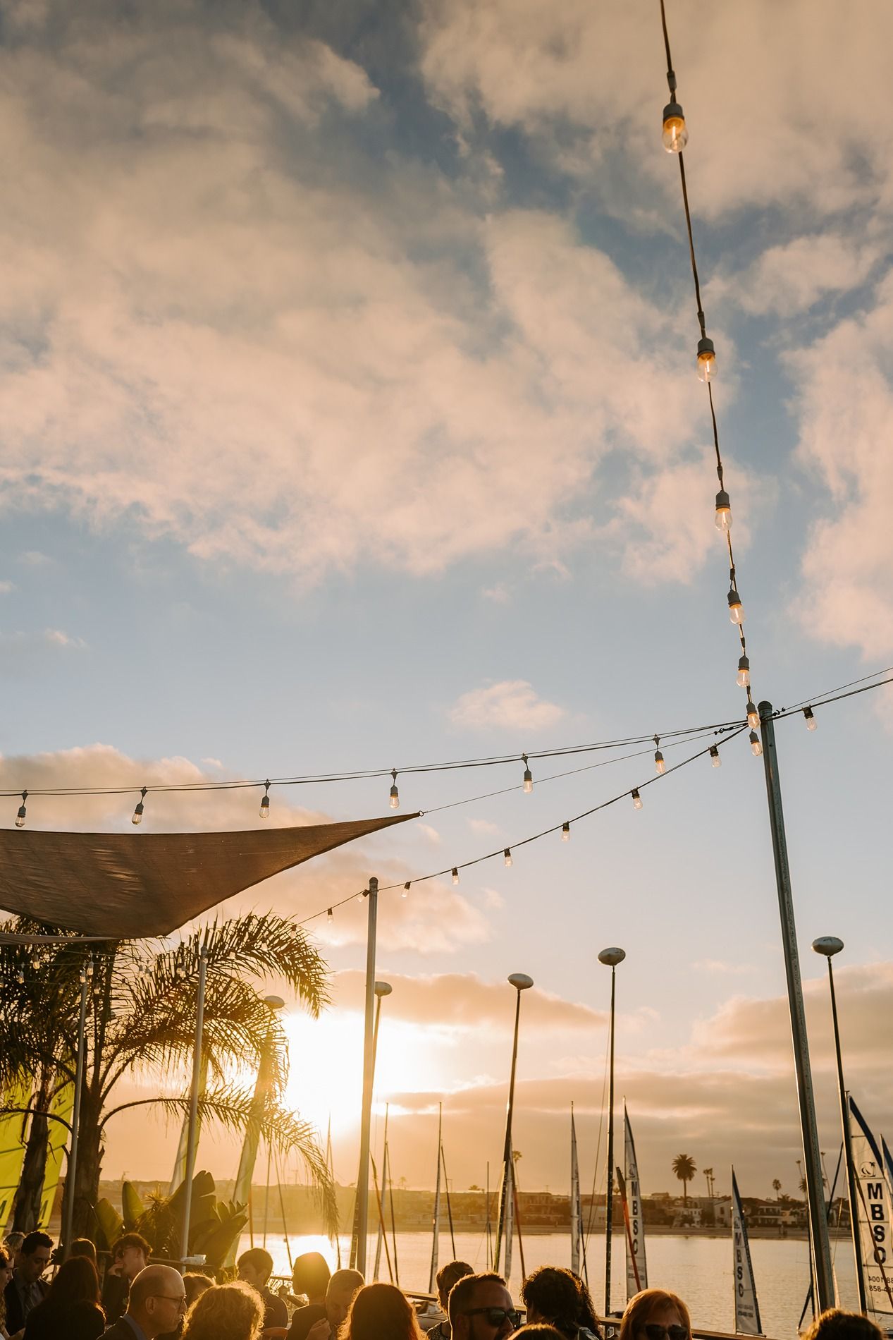Outdoor gathering at sunset with string lights, palm trees, and a waterfront view under a partly cloudy sky at Tower Beach Club.