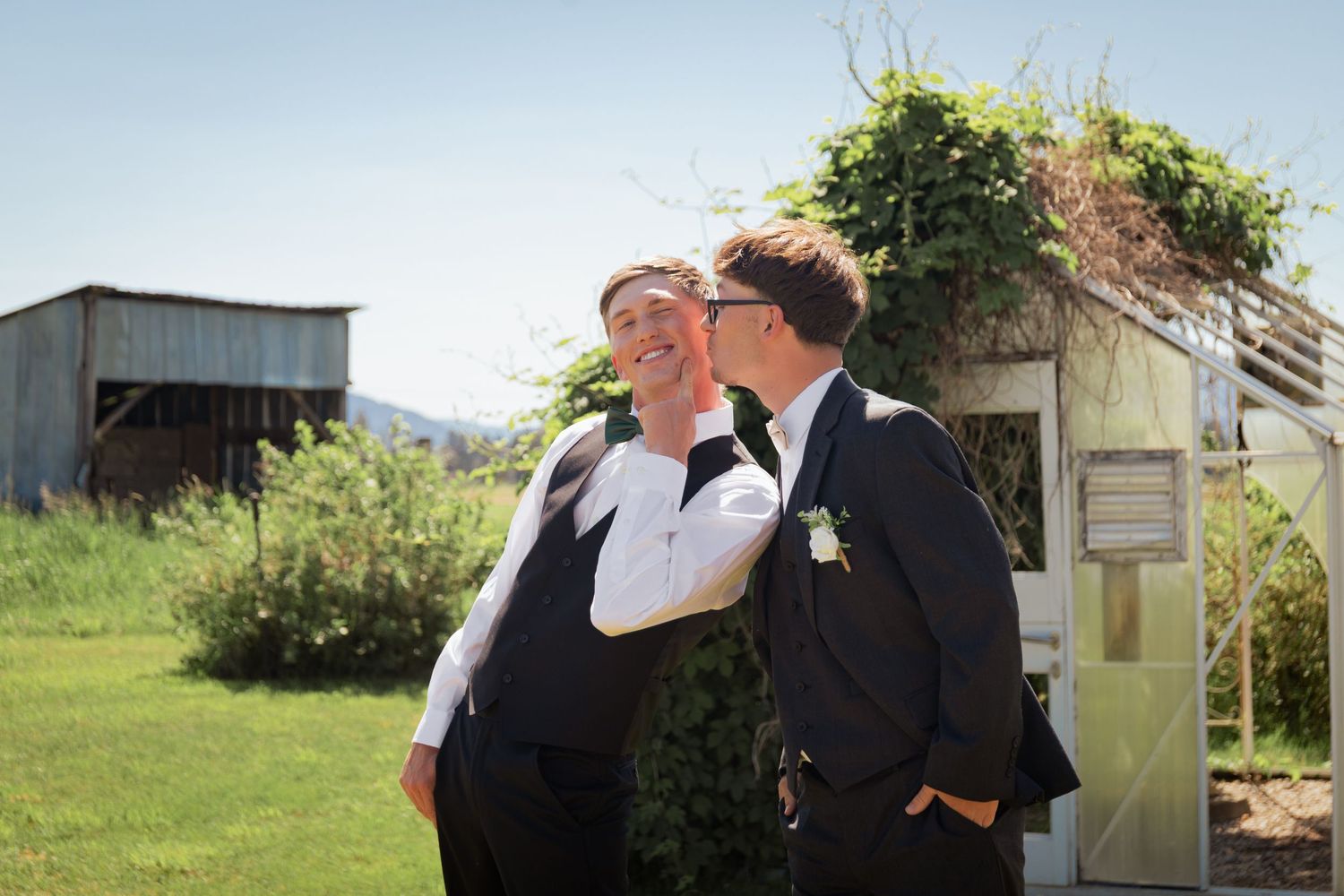 Two grooms share a tender kiss outdoors on their wedding day, with a rustic barn and greenery in the background.