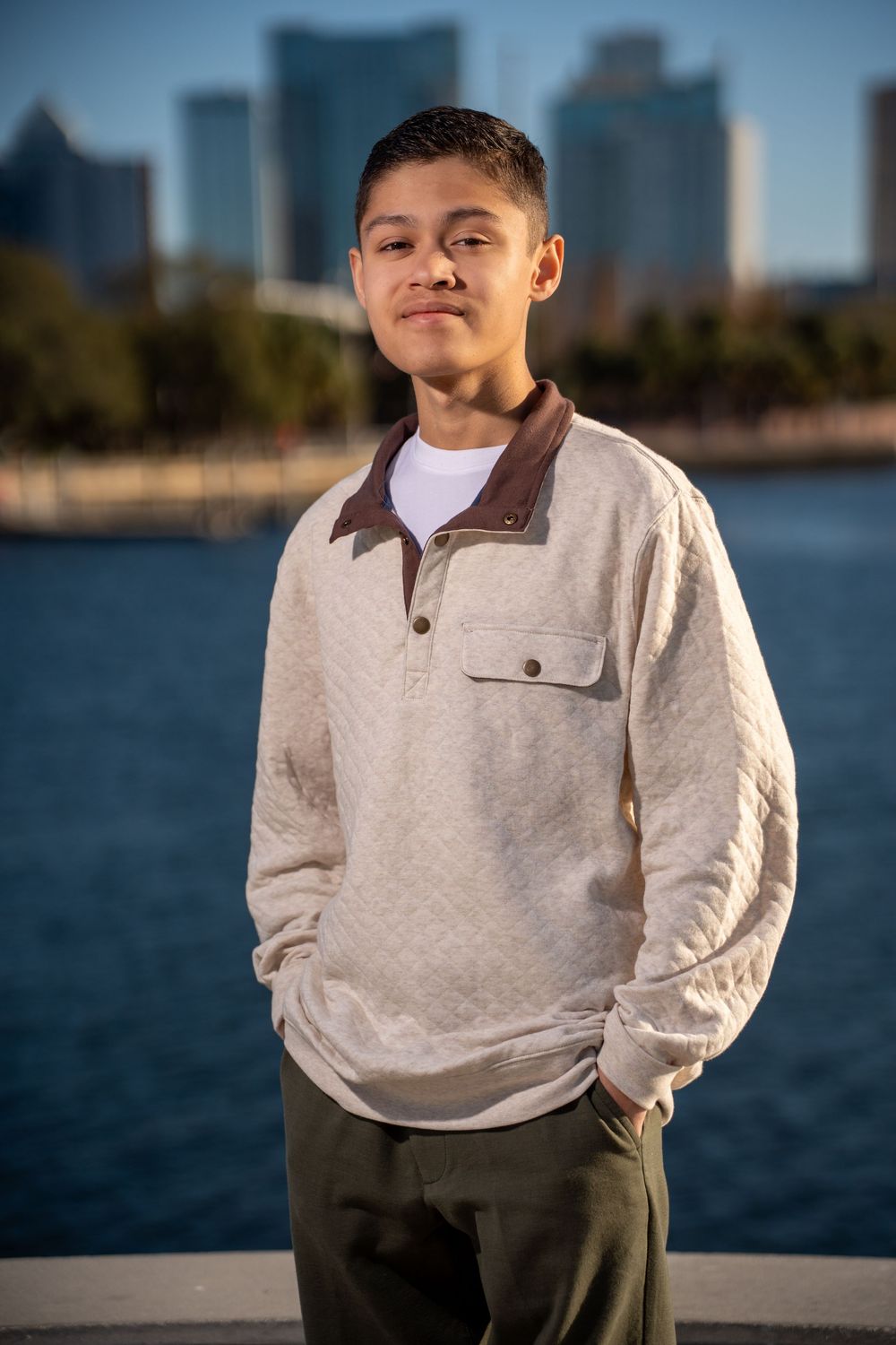 Young man in beige quilted pullover stands confidently by waterfront with city skyline backdrop.