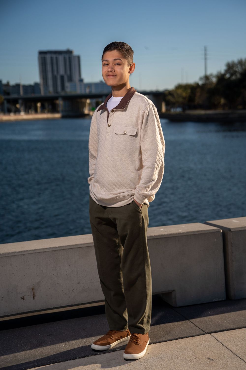 Young man in white pullover and olive pants standing by a urban waterway on a sunny day.