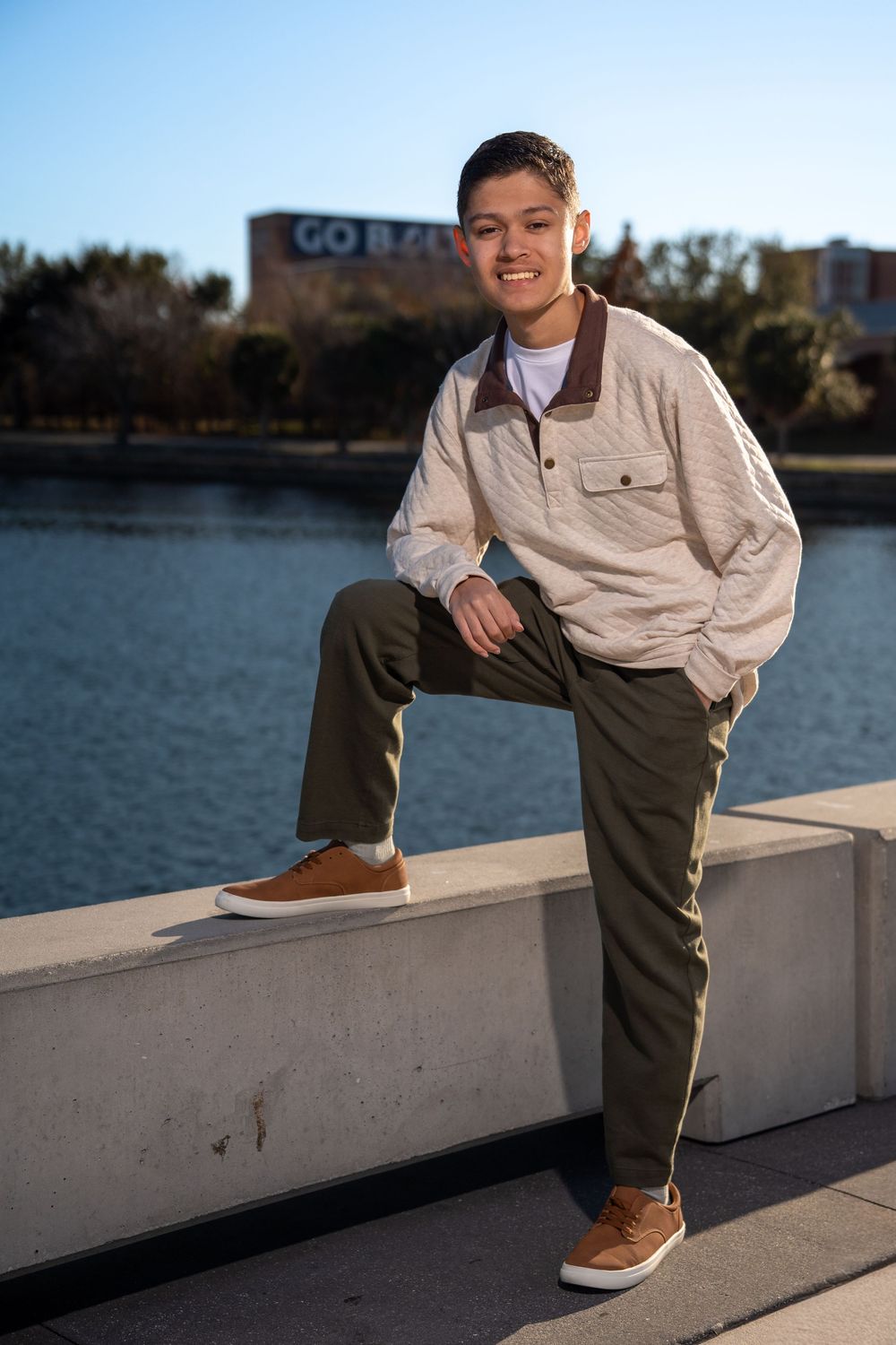 Young man smiling, seated on concrete ledge by a lake, wearing beige pullover and olive pants, with a GO BAYLOR sign behind.