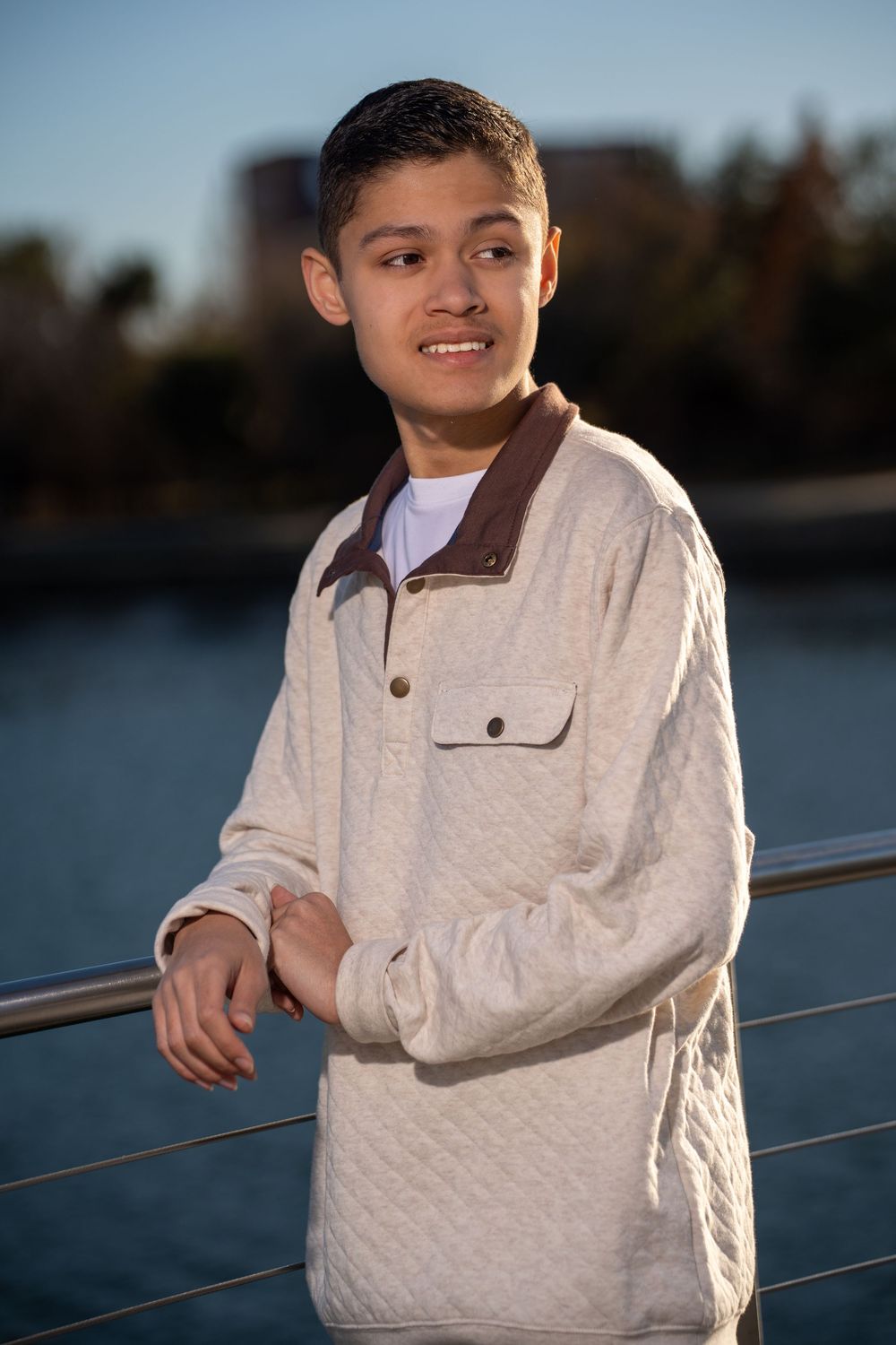 Smiling teen in beige quilted jacket leans on railing by sunlit waterfront with trees behind.