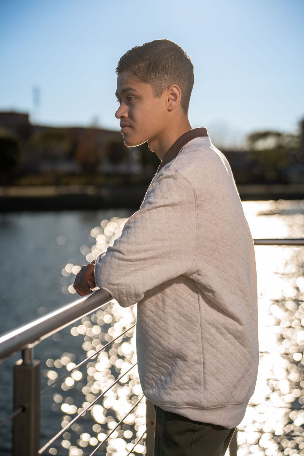 Young man in a gray quilted jacket leaning on a railing by a sunlit river, gazing thoughtfully into the distance.