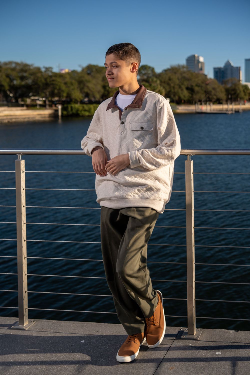 Young man in cream pullover and olive pants leaning on waterfront railing with city skyline backdrop.