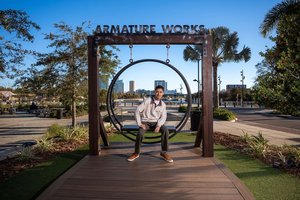 A man sits on a circular swing at Armature Works in Tampa, FL, with a waterfront skyline visible in the background.
