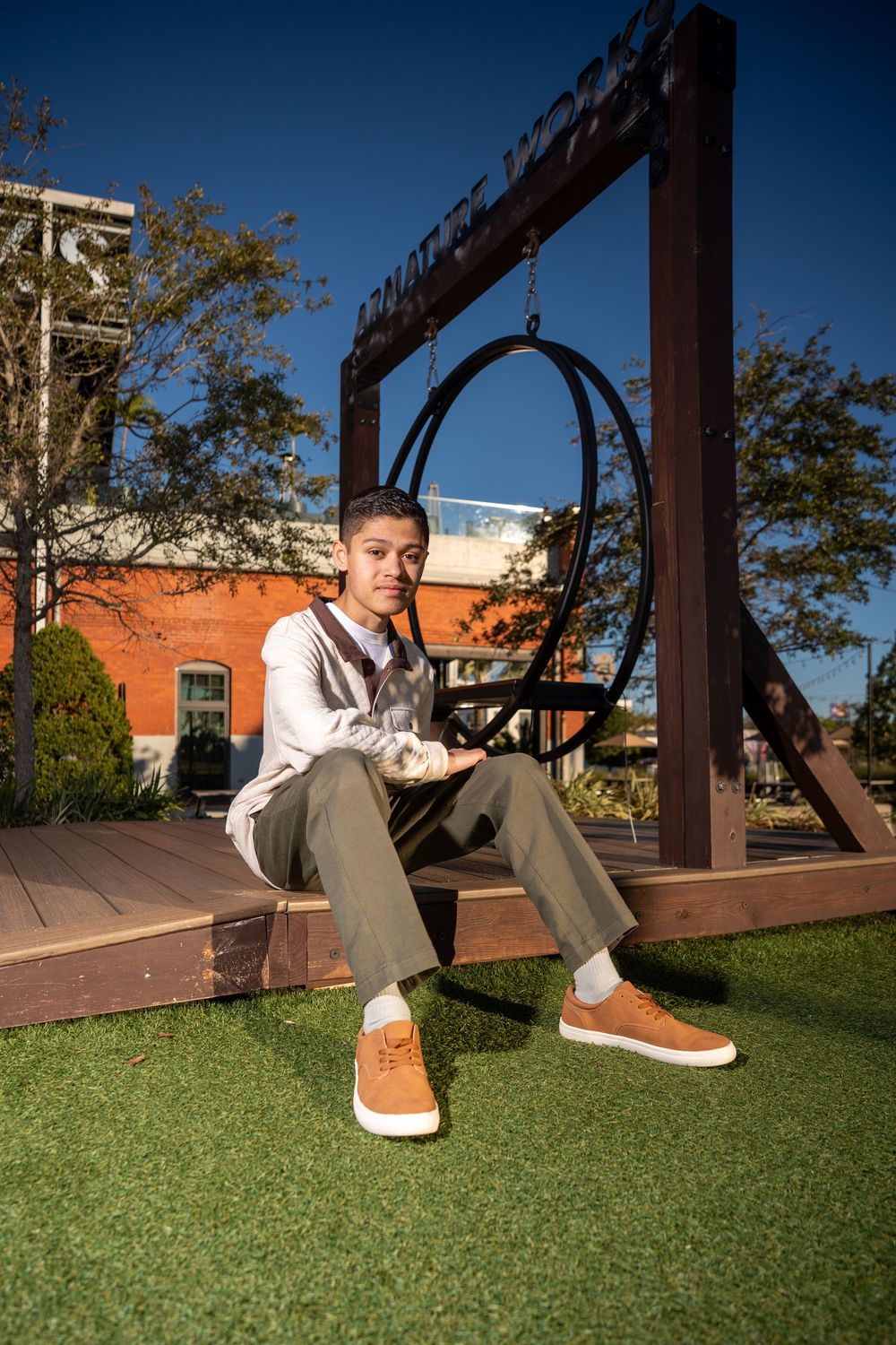 Young man sitting on wooden deck by circular swing, wearing khaki pants and tan sneakers outdoors.