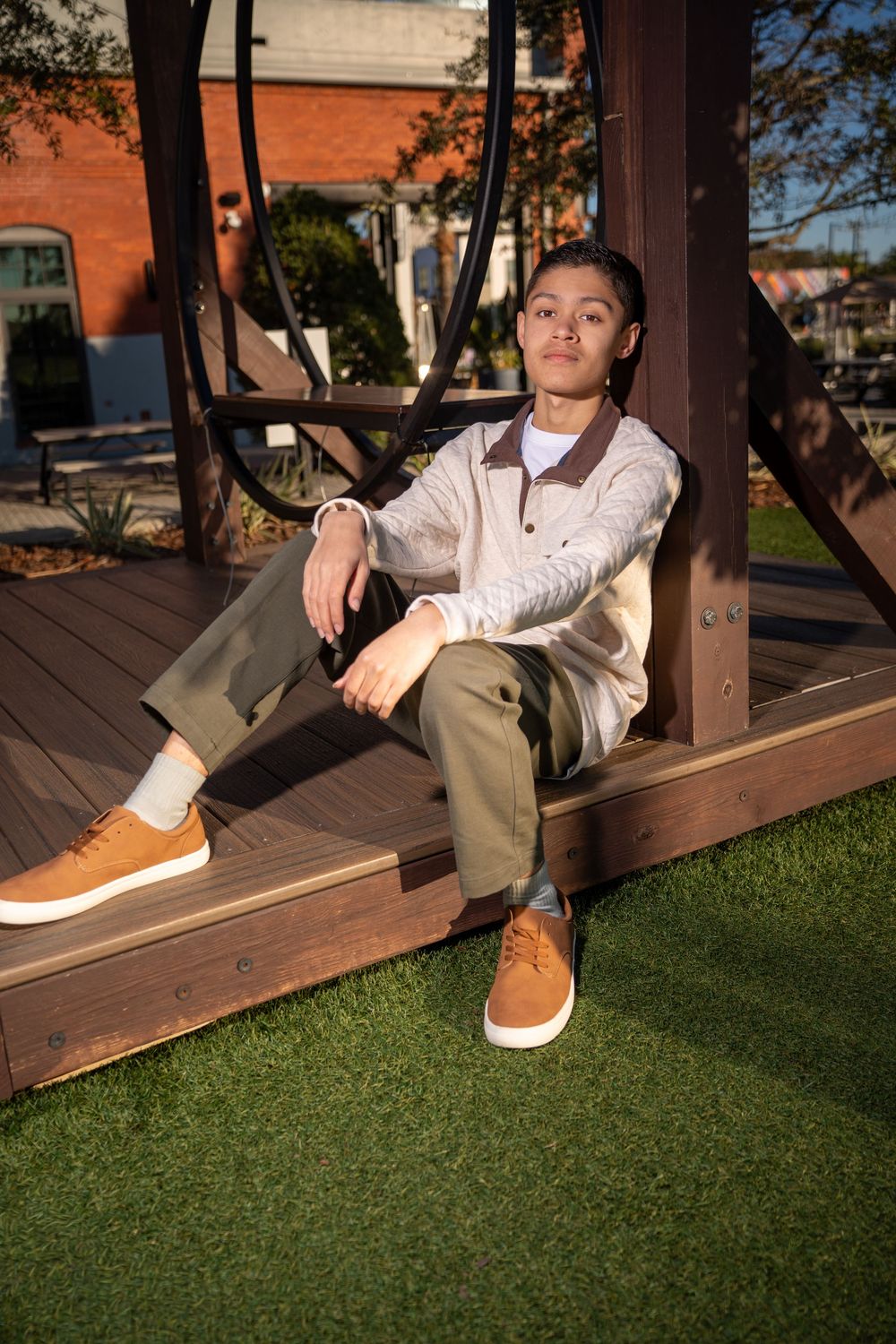 Young man in olive pants and tan sneakers sits relaxed on a wooden platform in a sunny outdoor urban setting.