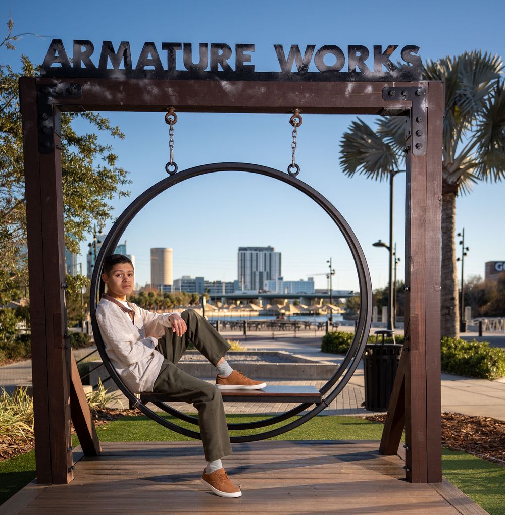 Woman sitting in a circular swing at Armature Works in Tampa, FL, with city skyline and palm trees in background.