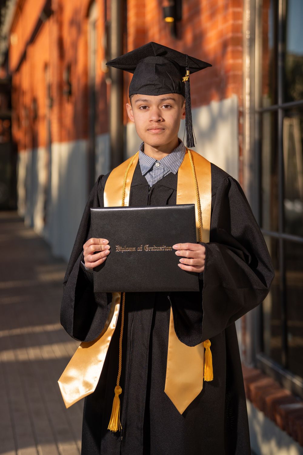 Young graduate in black cap and gown holding diploma, wearing gold honor stole, standing outdoors near brick building.