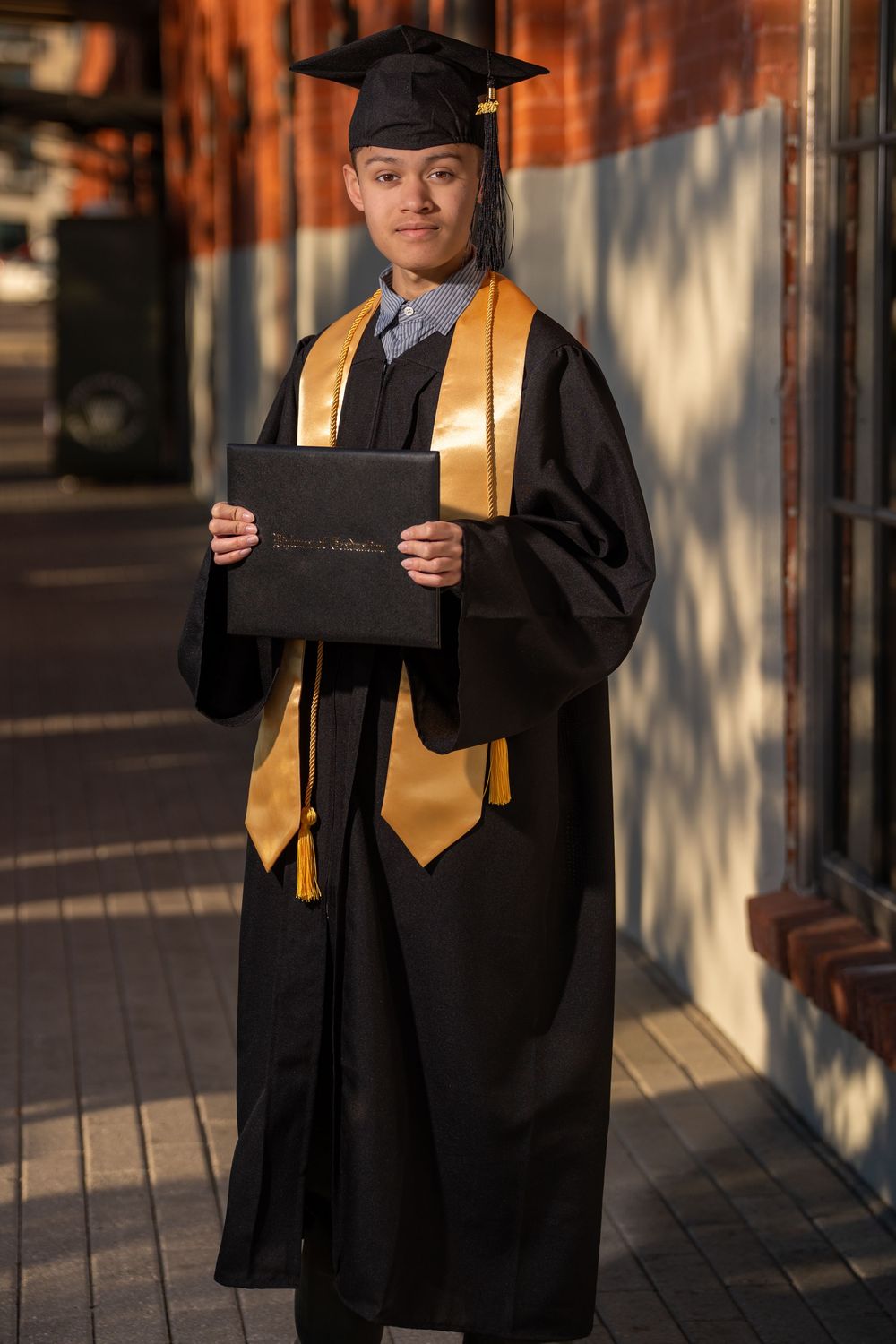 Young graduate in black and gold cap and gown holding diploma outdoors in sunlight.