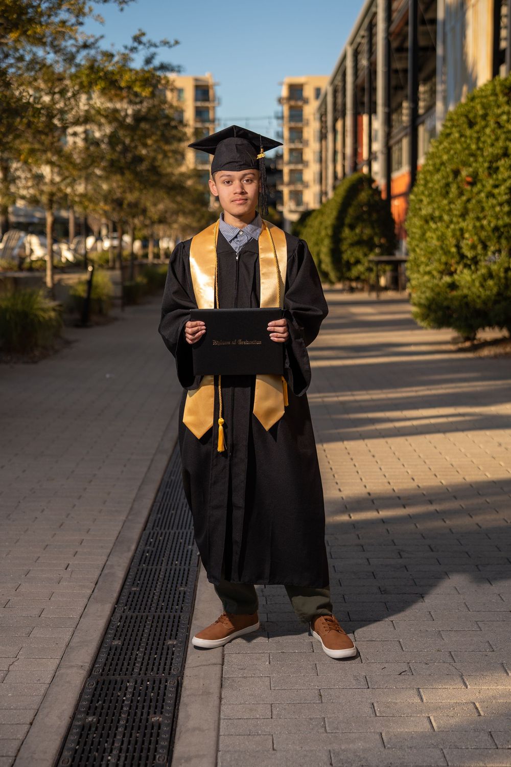 Graduate in black cap and gown with gold stole holds diploma on campus walkway.