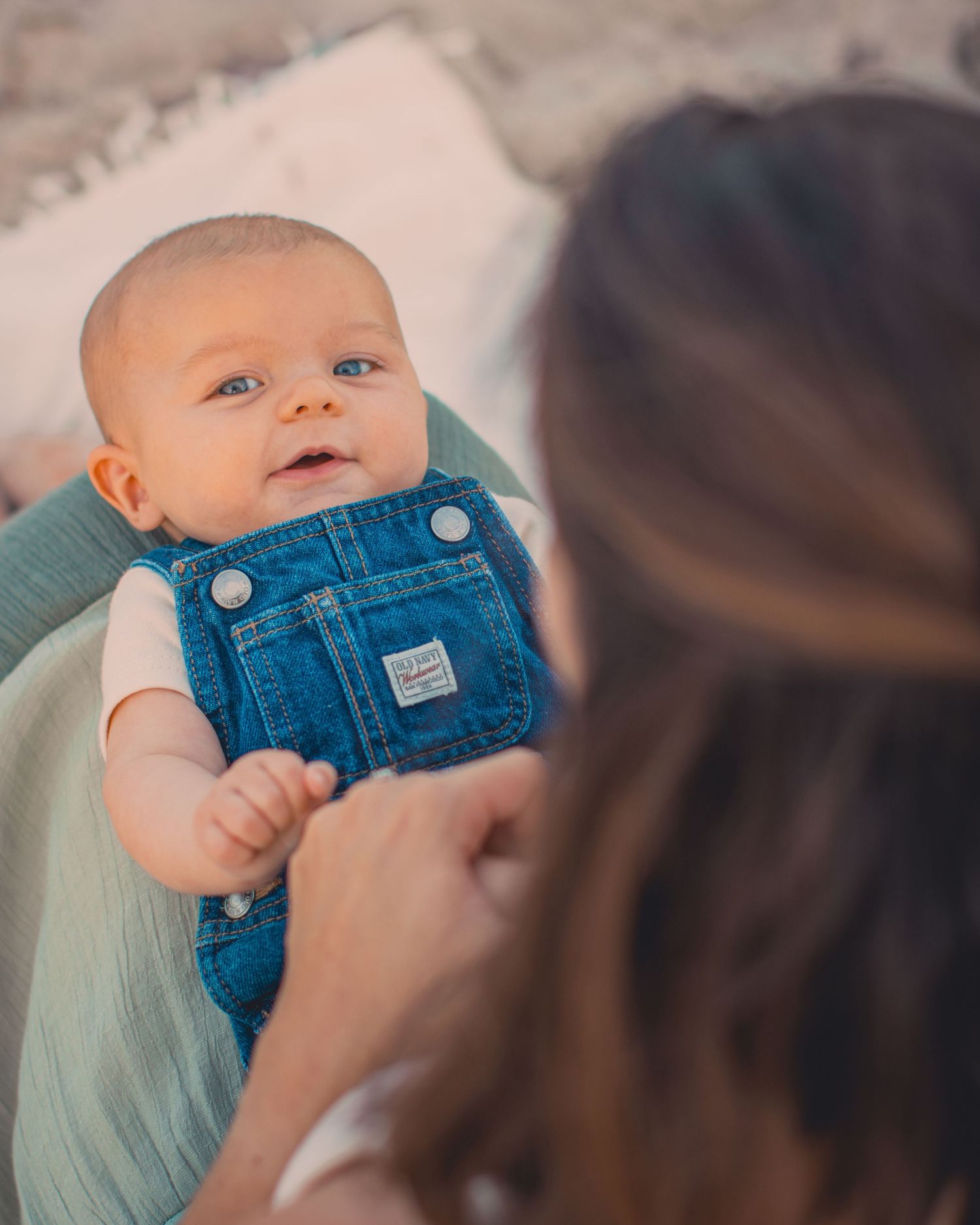 A smiling baby in denim overalls being held by a woman with brown hair during a tender moment.