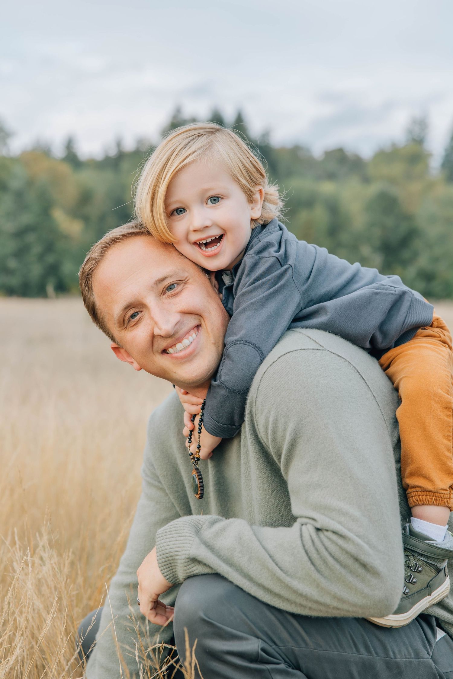 Smiling father giving laughing young child a piggyback ride in an open grassy field.